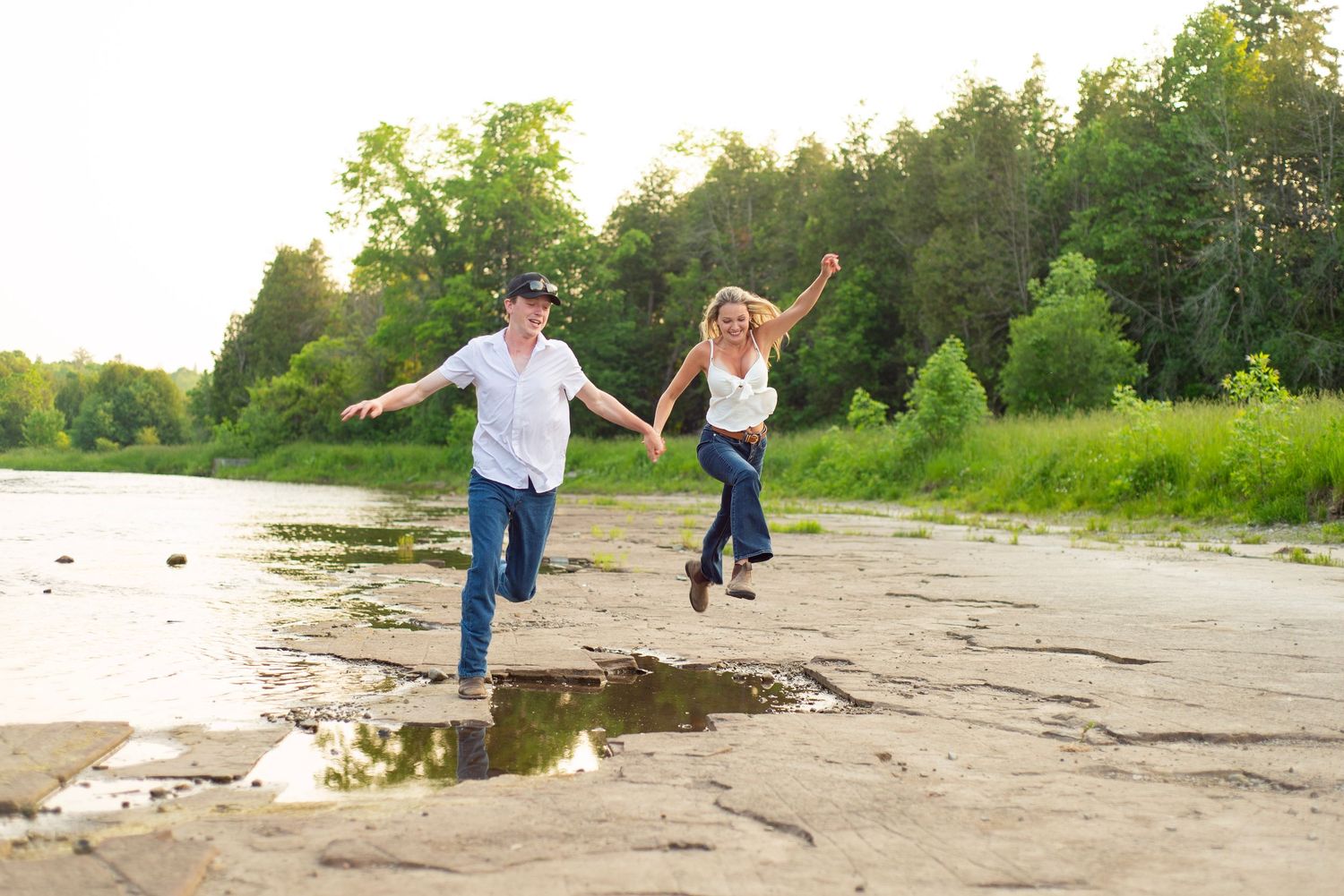 Two people hold hands and skip together on a rocky beach shoreline at sunset.