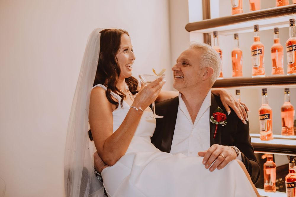 A couple smiles and embraces at what appears to be a restaurant bar with orange bottles in the background.