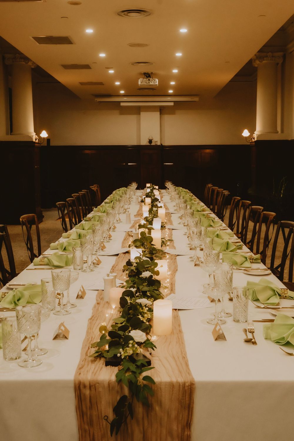 Long banquet tables with white linens and green place settings are decorated with greenery runners in an elegant room.