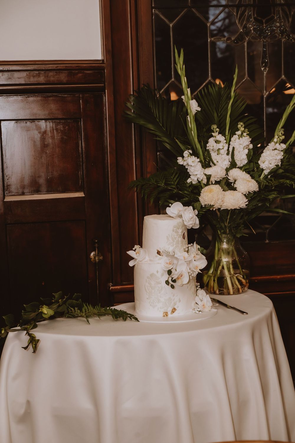 A stunning white wedding cake with floral decorations sits on a round table with elegant white tablecloth.