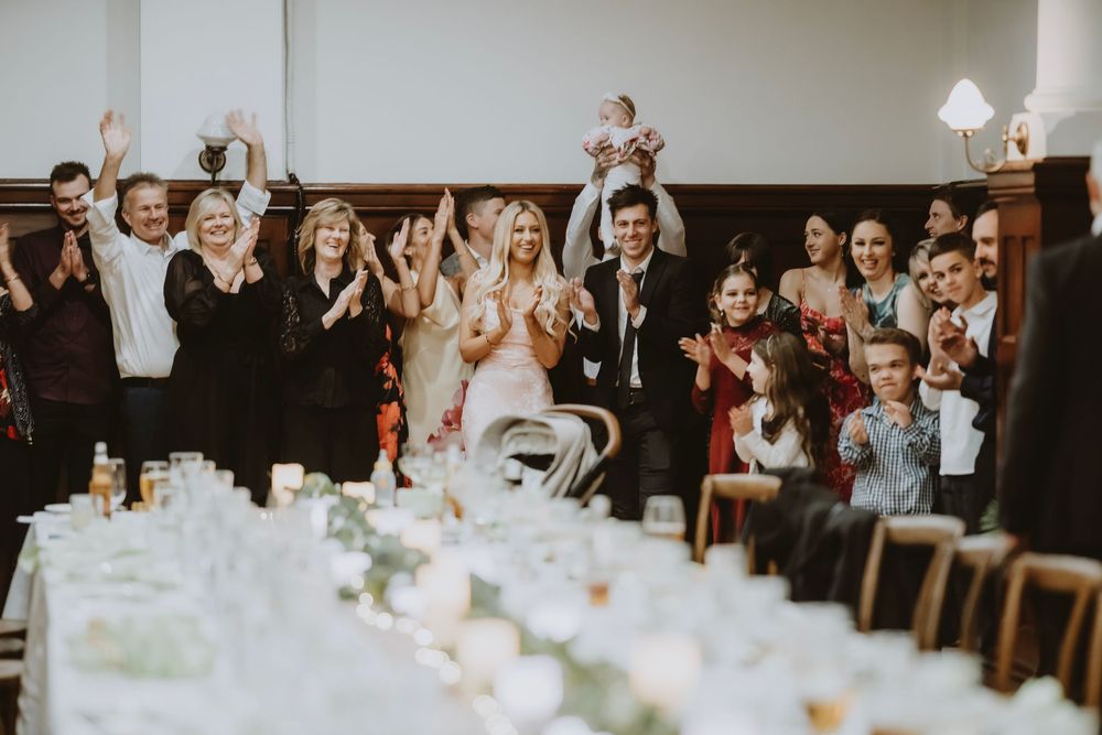 Wedding reception guests raise their arms and cheer during dinner celebration at long tables decorated in white.