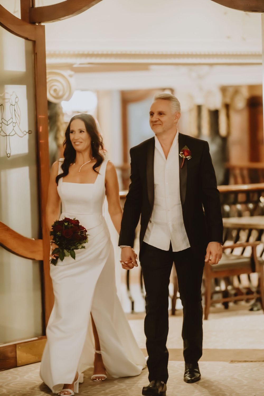 A couple in wedding attire walks together down a stylish hallway holding hands, captured in warm sepia tones.