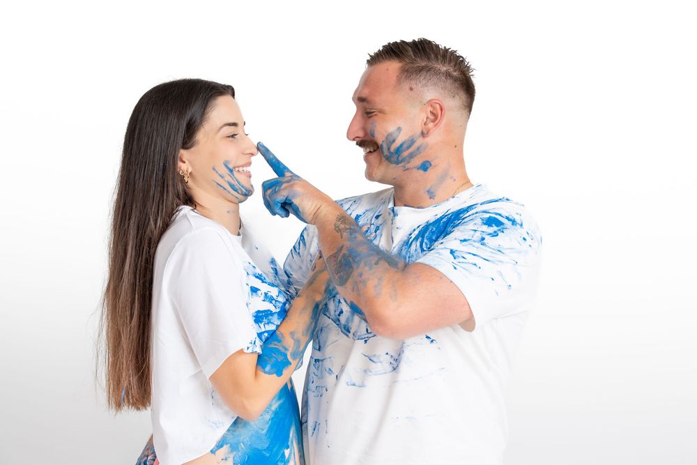 Playful couple with blue paint on their faces laughing and smiling during a creative photo session against white backdrop.