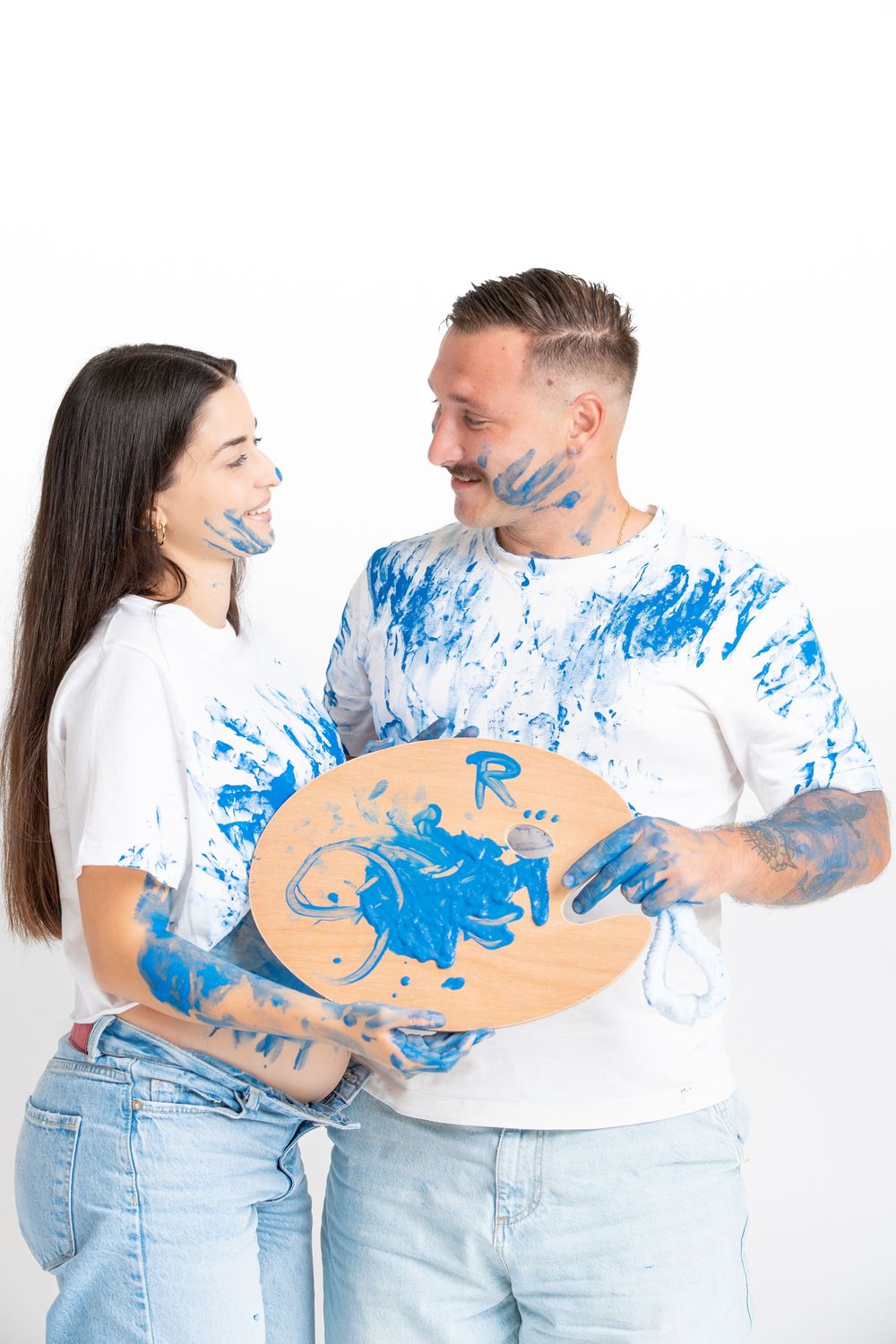 A couple in white t-shirts and jeans playfully creating a gender reveal paint splatter in blue paint.