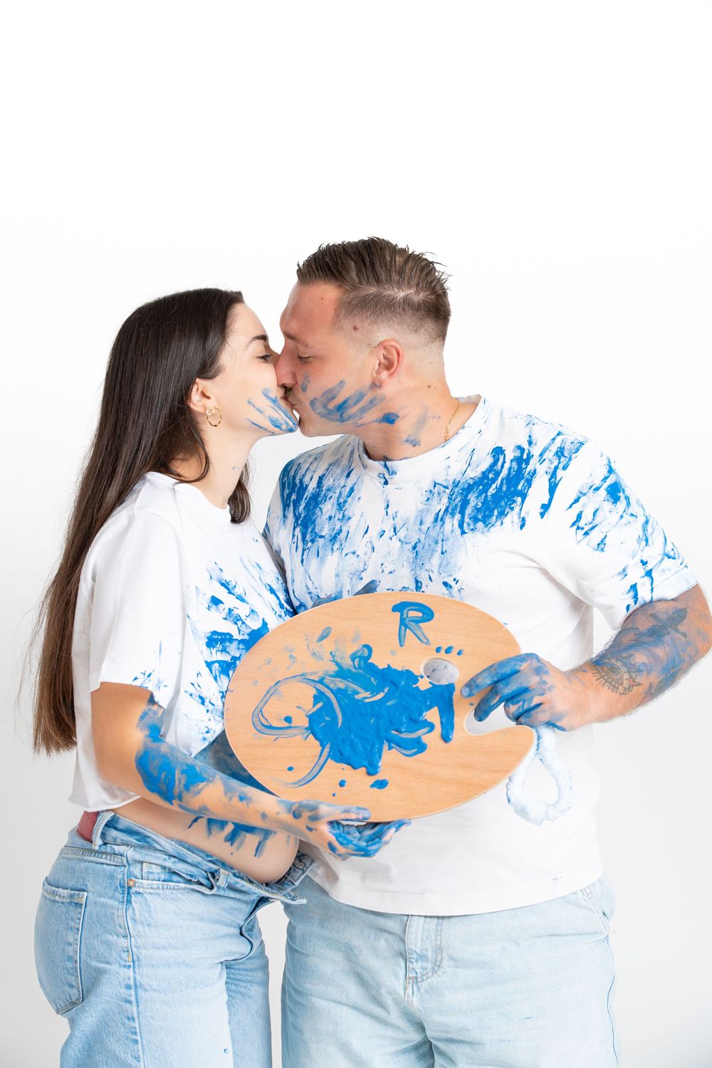 A couple in white shirts and denim jeans embrace while holding paint-covered palettes with blue splatters.