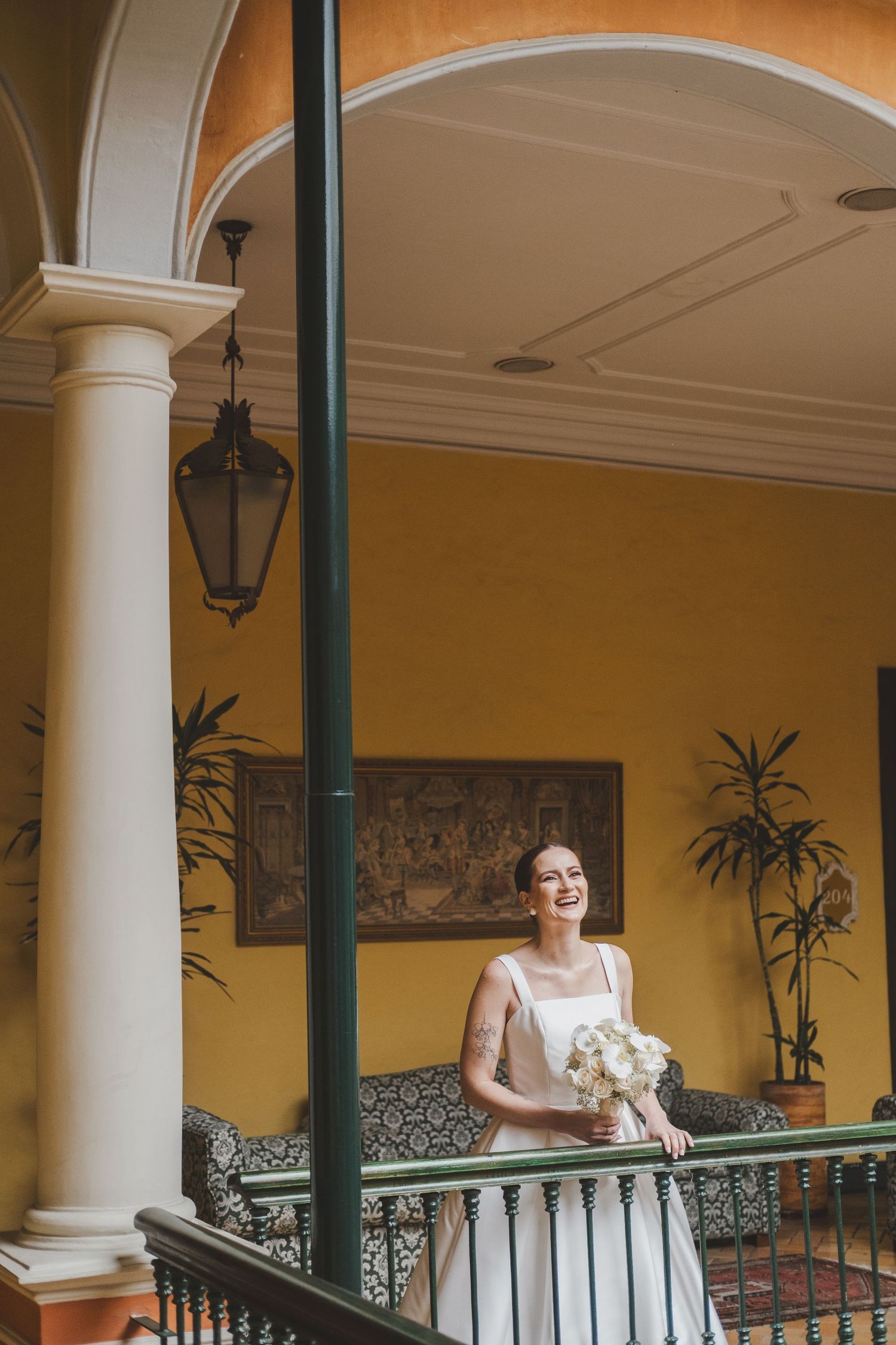 Elegant person in light colored dress standing on a covered balcony with ornate railings and yellow walls.