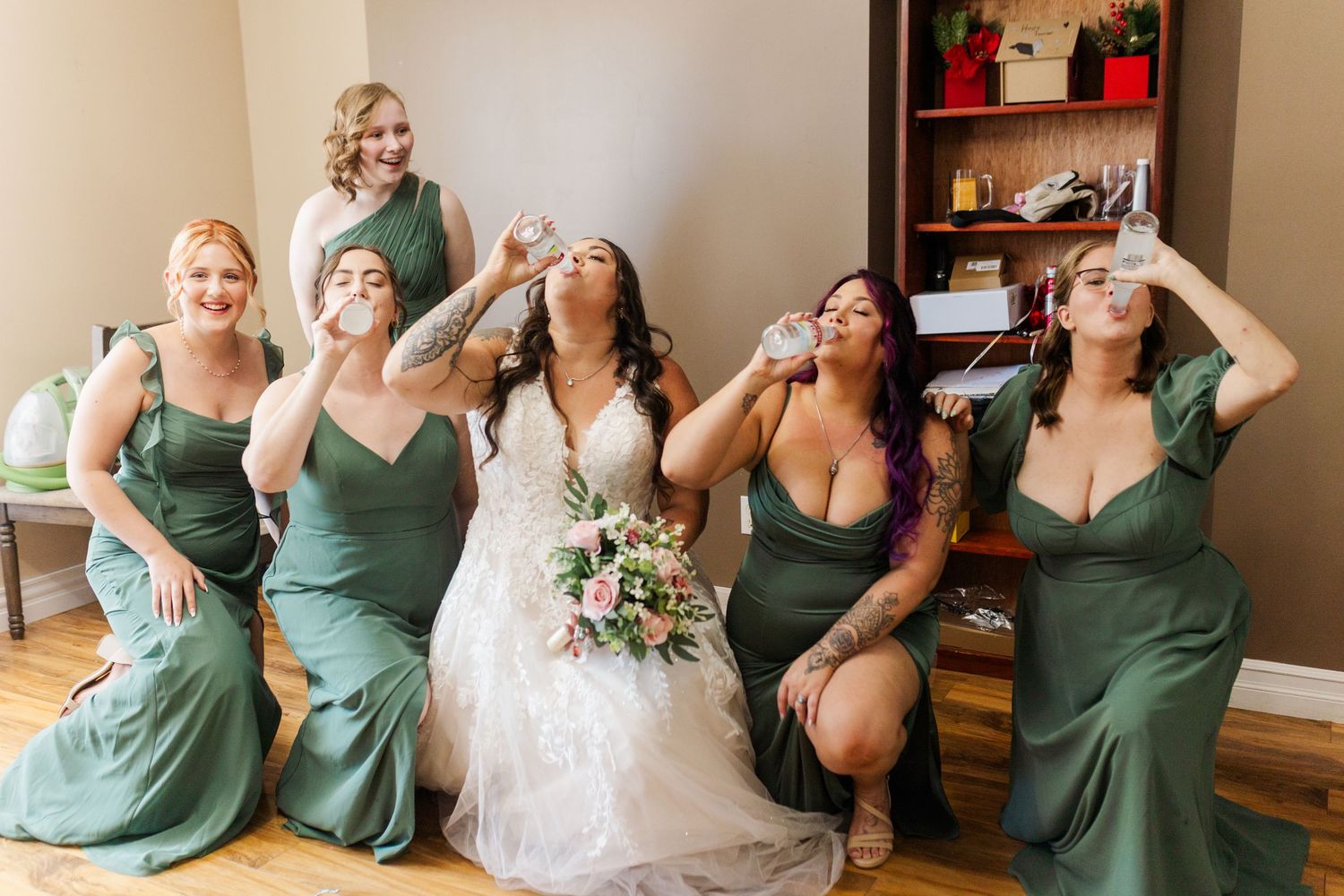 Wedding party in emerald green dresses gather together with white bouquet for a celebratory toast.