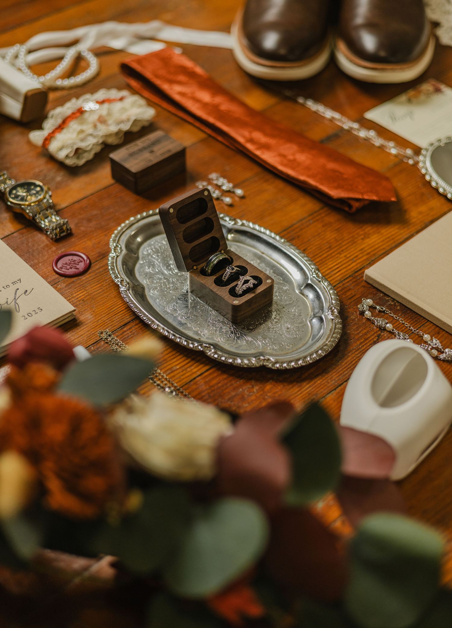 Wedding accessories including crystal sandals, rings in wooden box, floral petals and decorative elements on rustic wood surface.