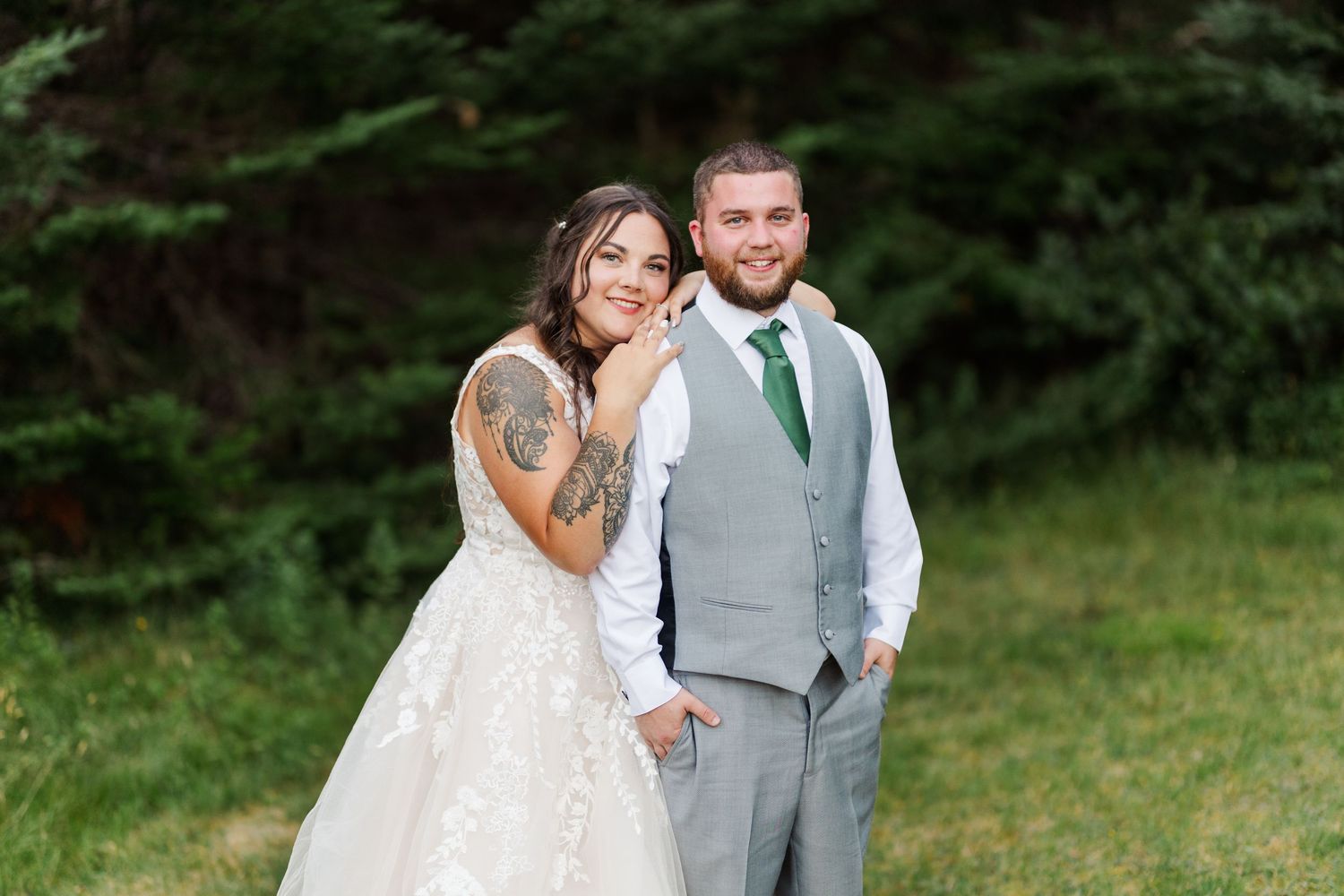 A couple in wedding attire pose together outdoors against evergreen trees during a summer celebration.