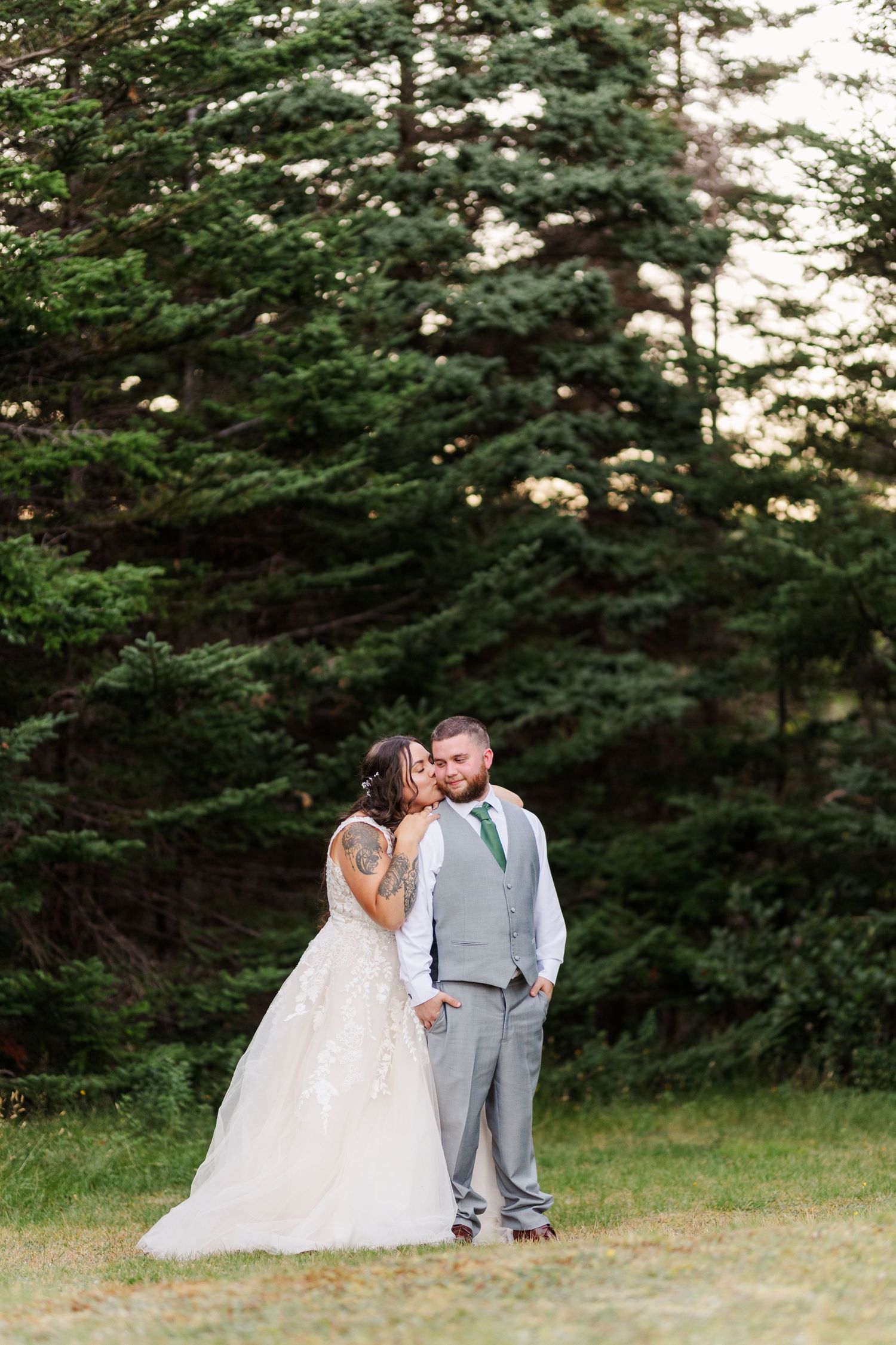 A couple shares an intimate moment in their wedding attire against a backdrop of tall evergreen trees in a forest clearing.