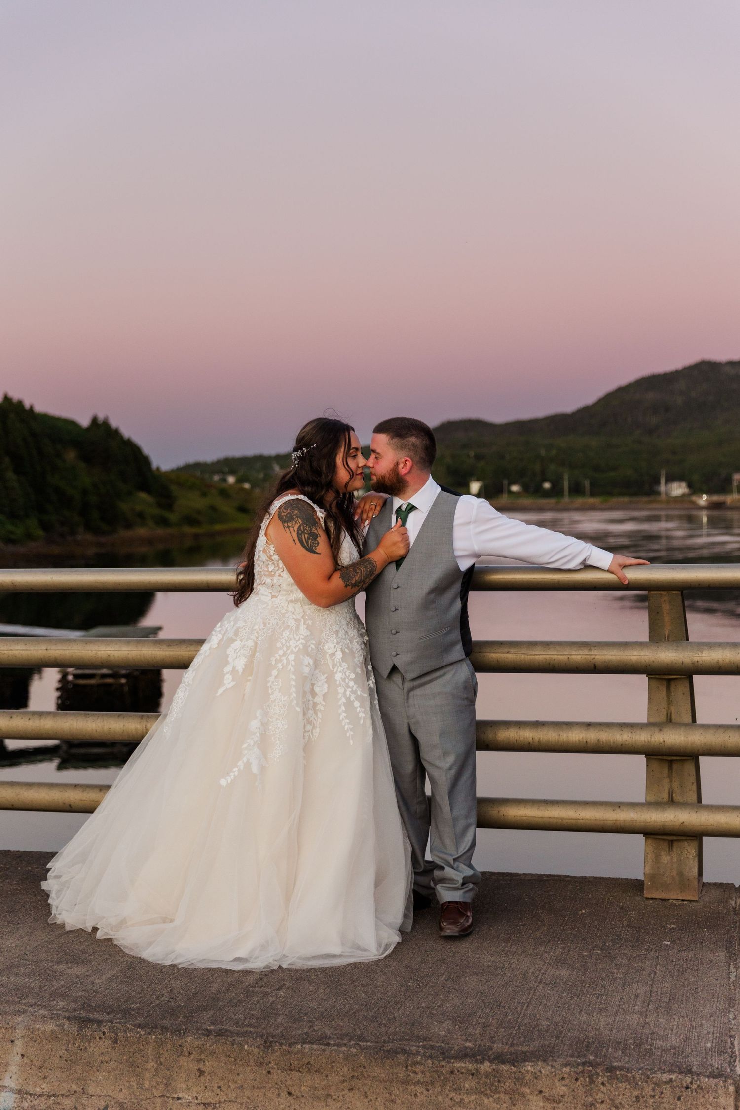 A romantic wedding photo showing a couple embracing on a bridge at sunset with mountains in the background.
