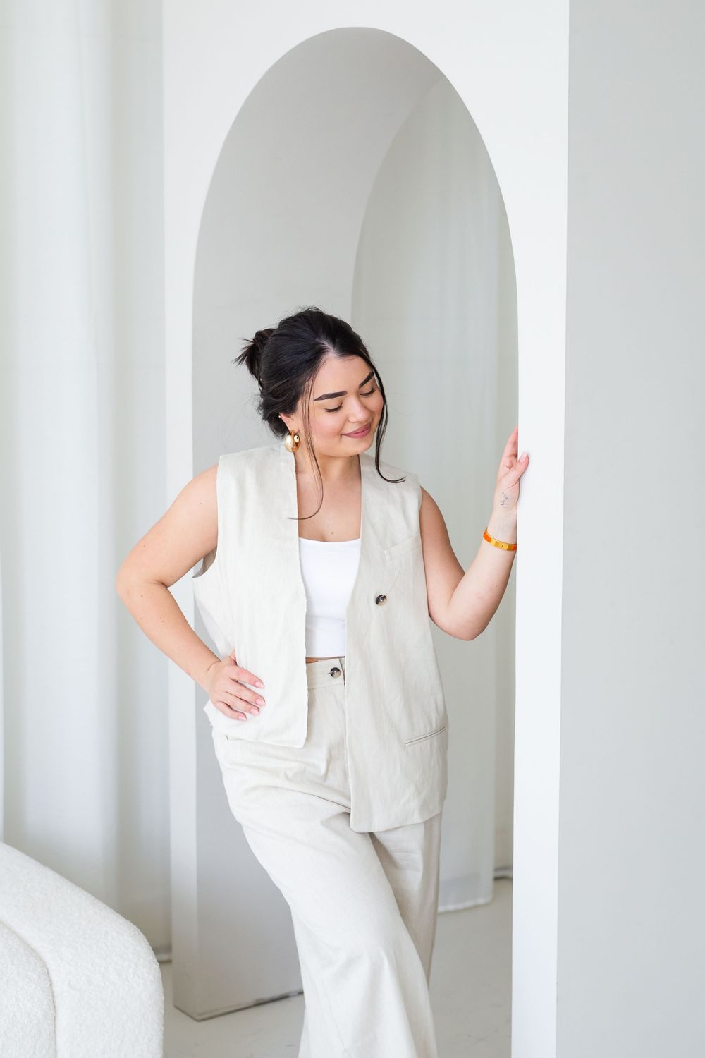 Figure in a cream-colored linen vest and pants ensemble poses against a white archway in a minimalist setting.