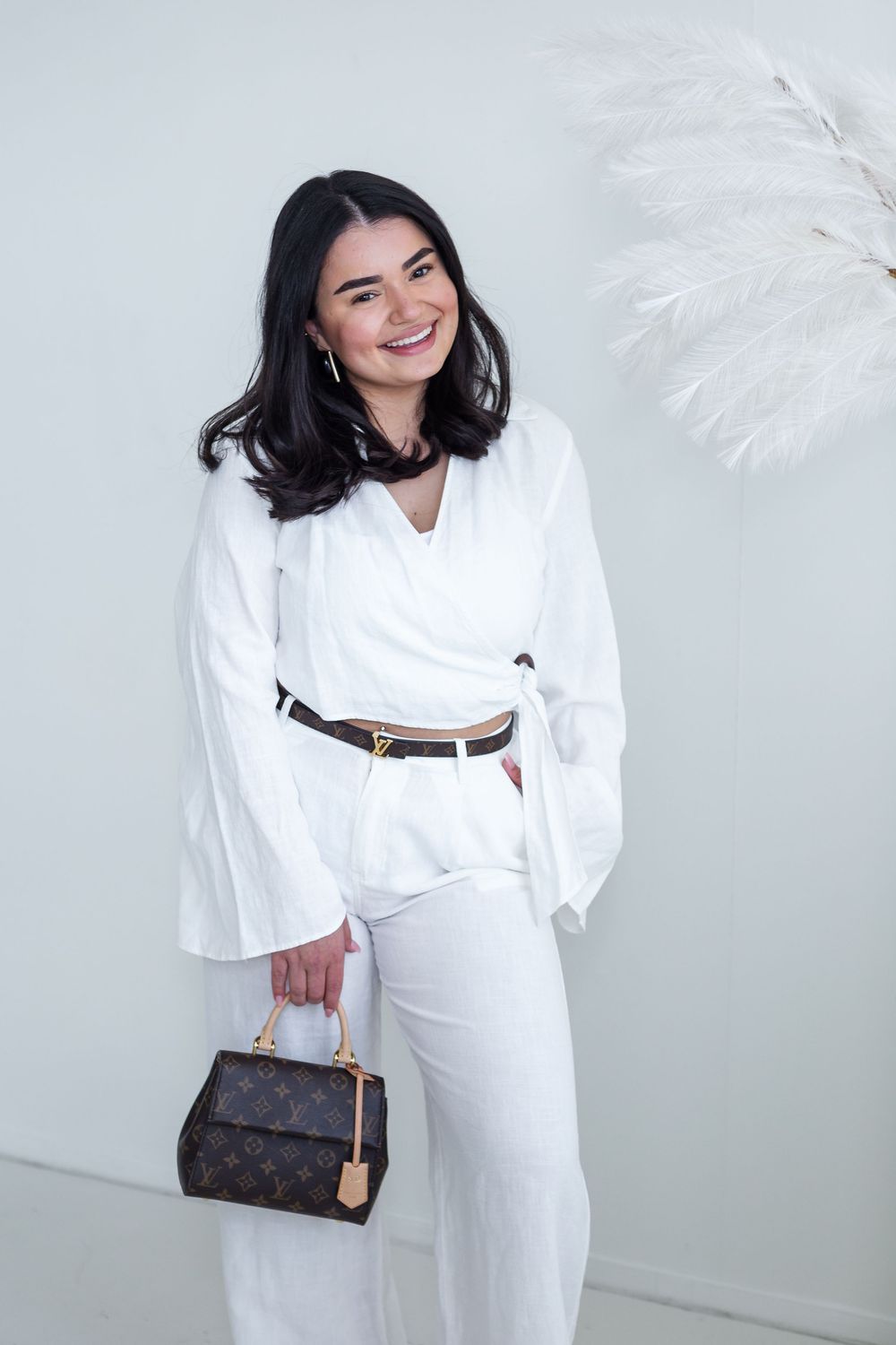 A chic all-white outfit featuring a wrapped top with bell sleeves, wide-leg pants, brown belt, and Louis Vuitton handbag.