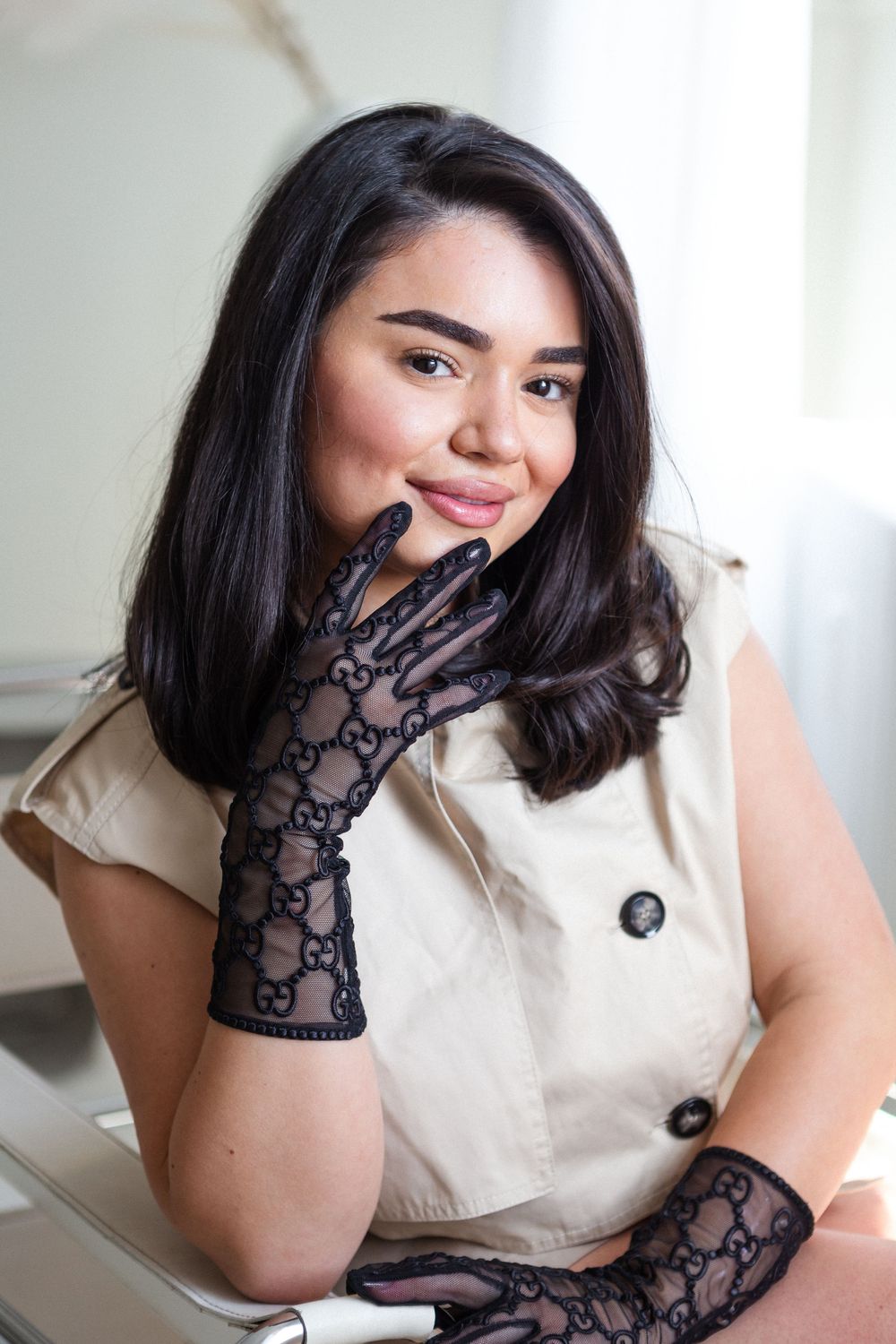 Woman in cream colored sleeveless top poses wearing elegant black lace gloves with hand near face.