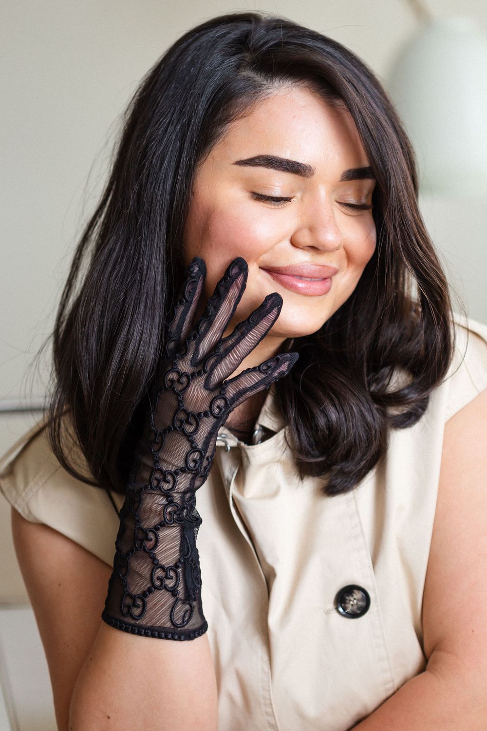 Person wearing stylish black lace gloves poses with eyes closed and gentle smile in cream-colored outfit.