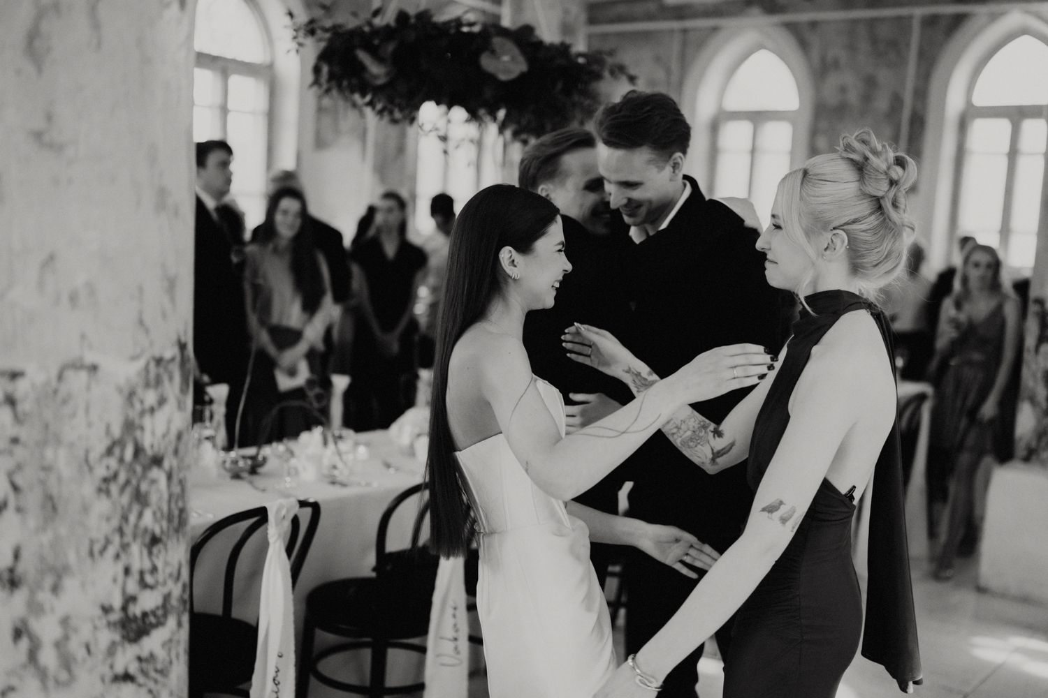 Elegant wedding reception scene with guests dancing in a historic venue with arched windows.