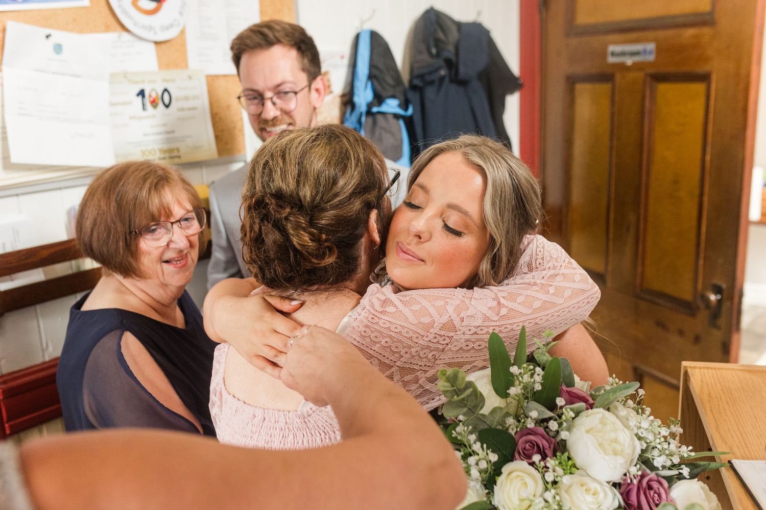 A tender embrace shared at a casual wedding celebration with flowers and well-wishers in the background.