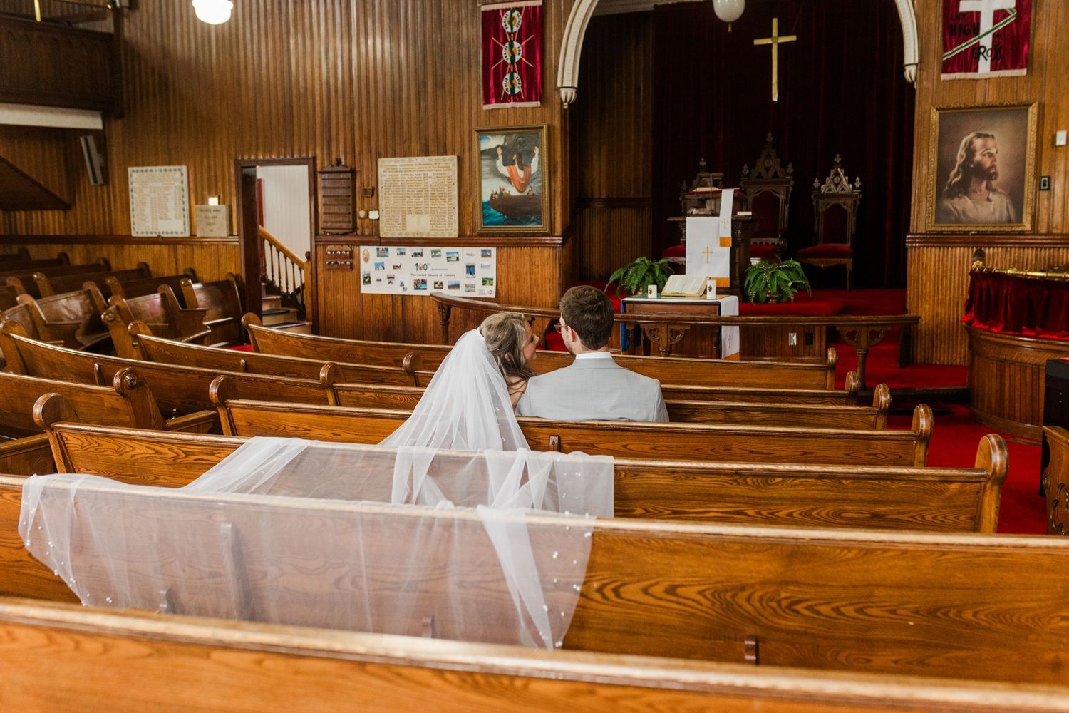 Two people sit alone in wooden church pews during an intimate ceremony, with one wearing a flowing white veil.