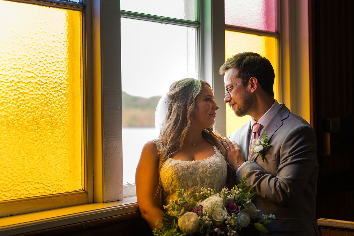 A couple shares an intimate moment by colorful stained glass windows while holding a bouquet of white and purple flowers.