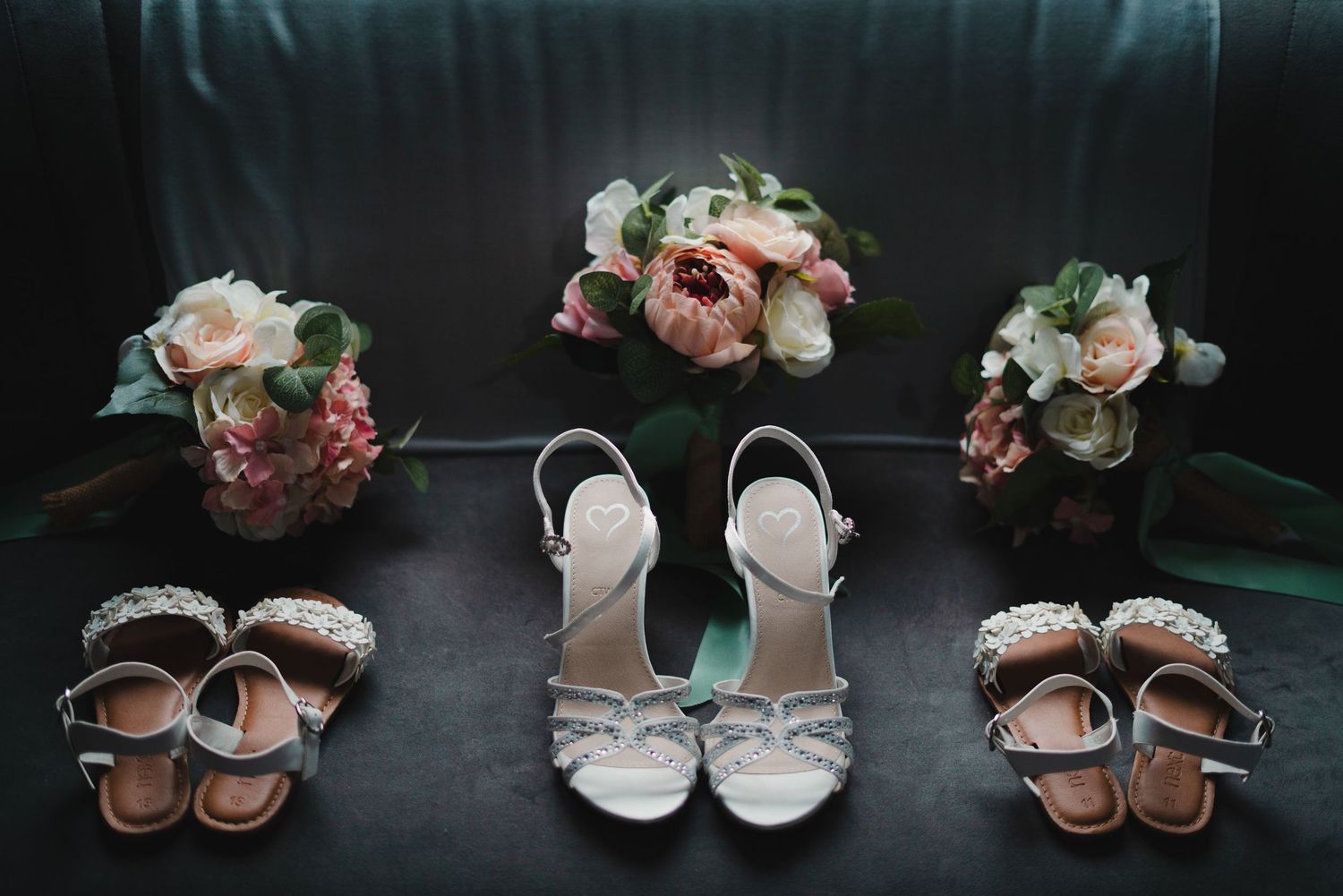 Collection of bridal shoes and sandals arranged with pink floral bouquets on dark background.