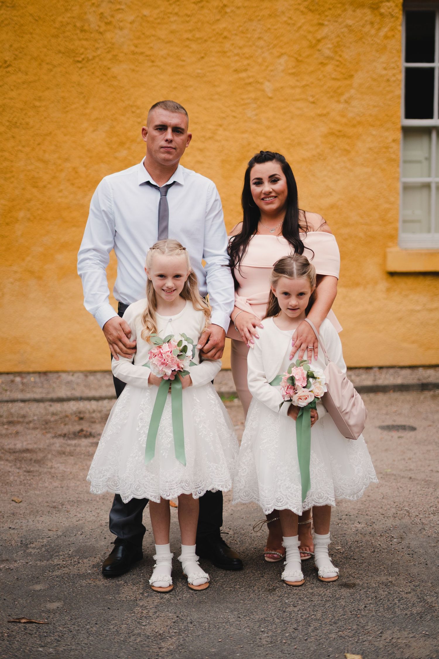 Family portrait with flower girls in white dresses holding small bouquets against yellow wall.