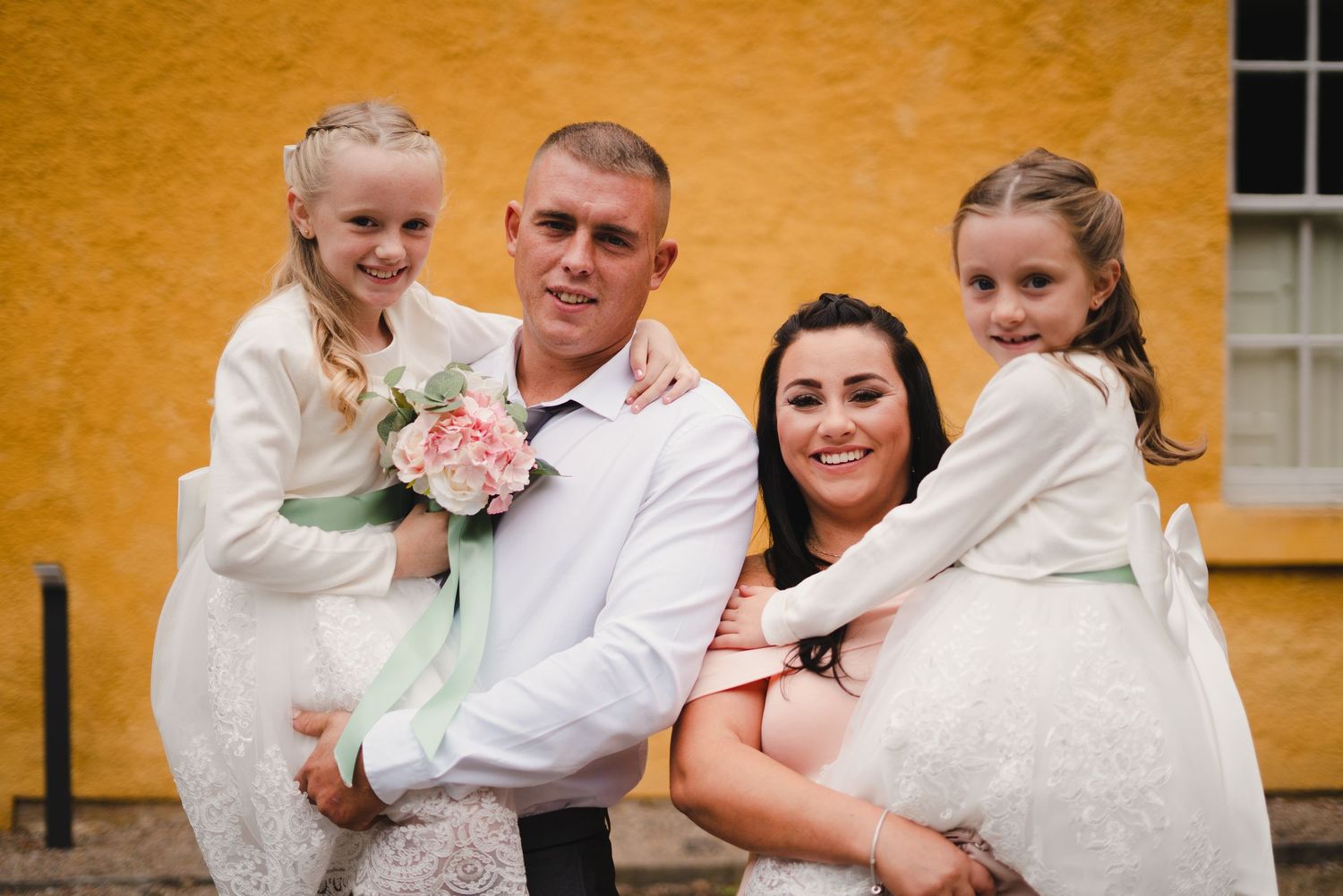 Wedding party posing outdoors against a yellow building wall.