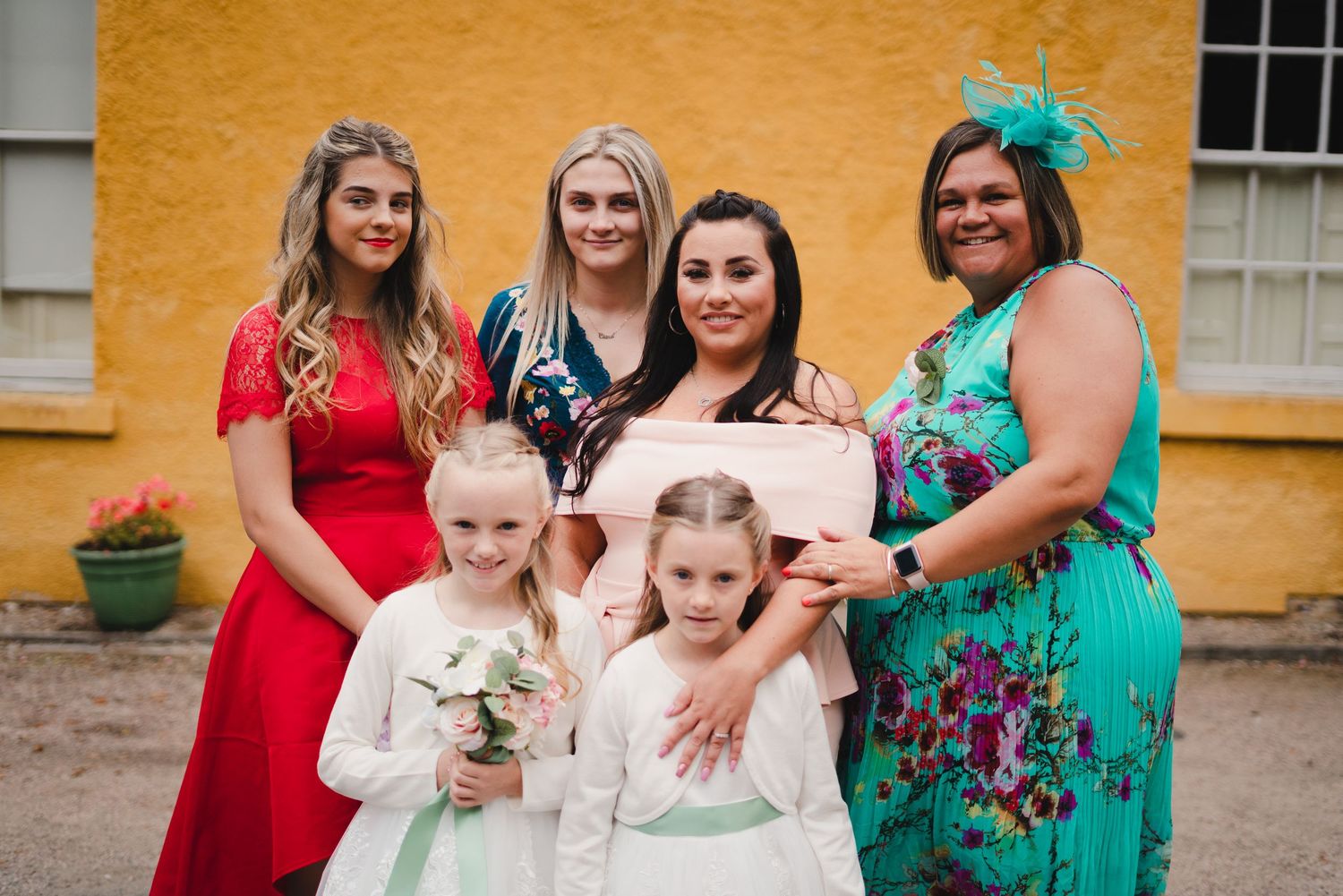 Group photo of wedding party members wearing colorful dresses against yellow building backdrop.