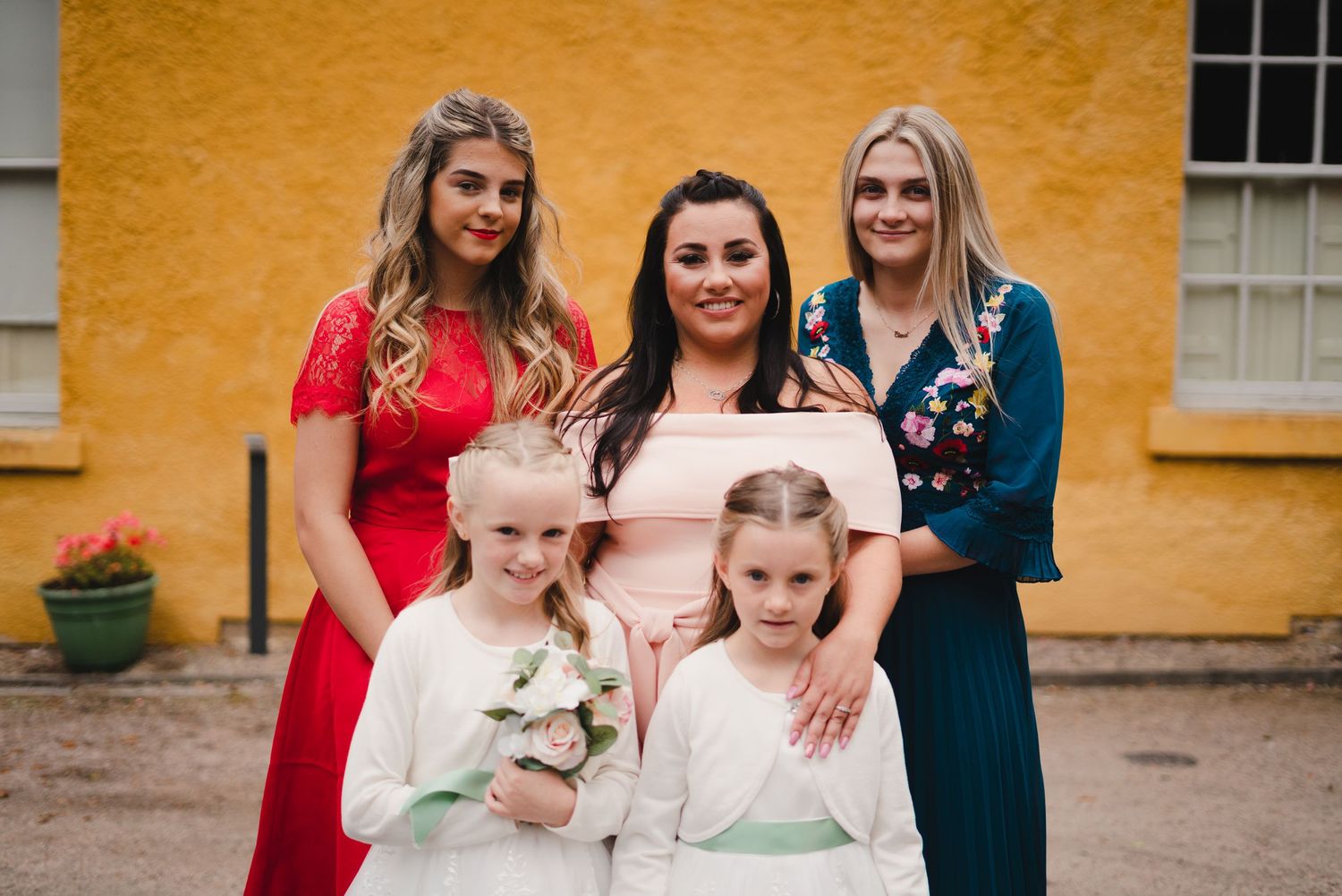Group photo against yellow wall featuring wedding attendees in colorful dresses.