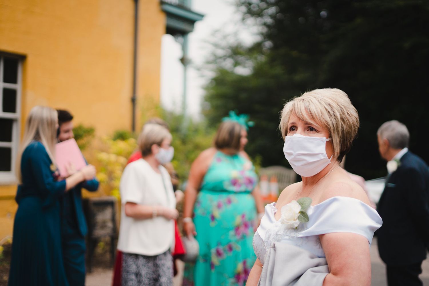 Guests socializing at an outdoor event near a yellow building while wearing protective masks.