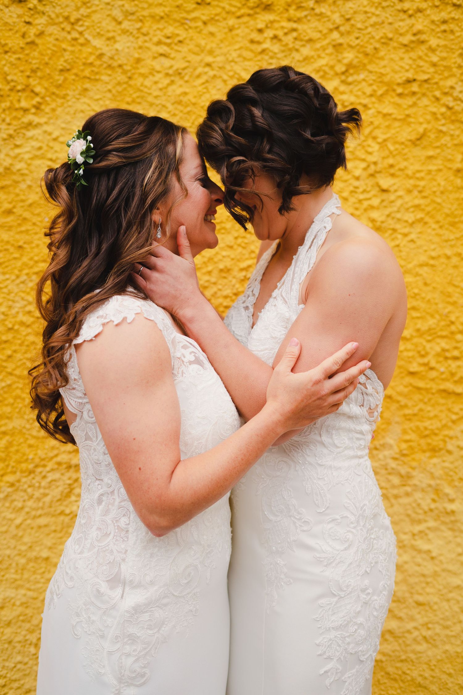 Couple in white lace wedding dresses share a romantic moment against vibrant yellow wall.