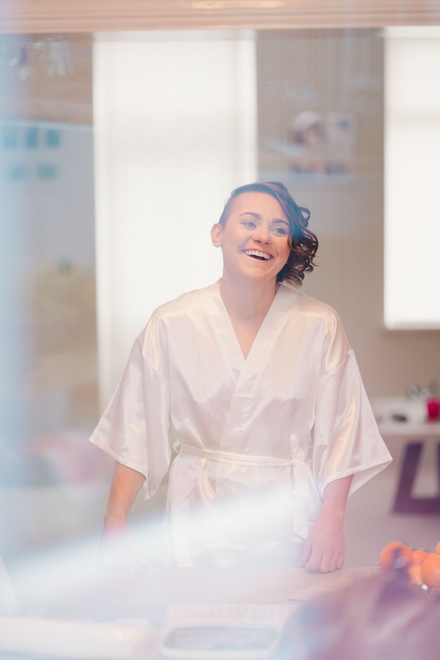 Person in white robe laughs joyfully in bright sunlit room during wedding preparations.