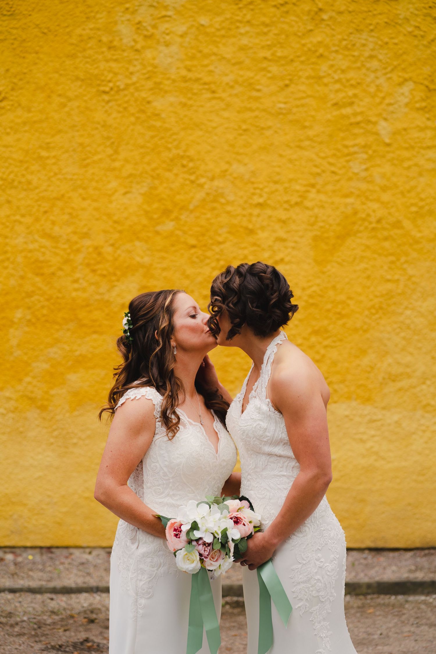 A romantic moment between two people in white dresses against a vibrant yellow wall.