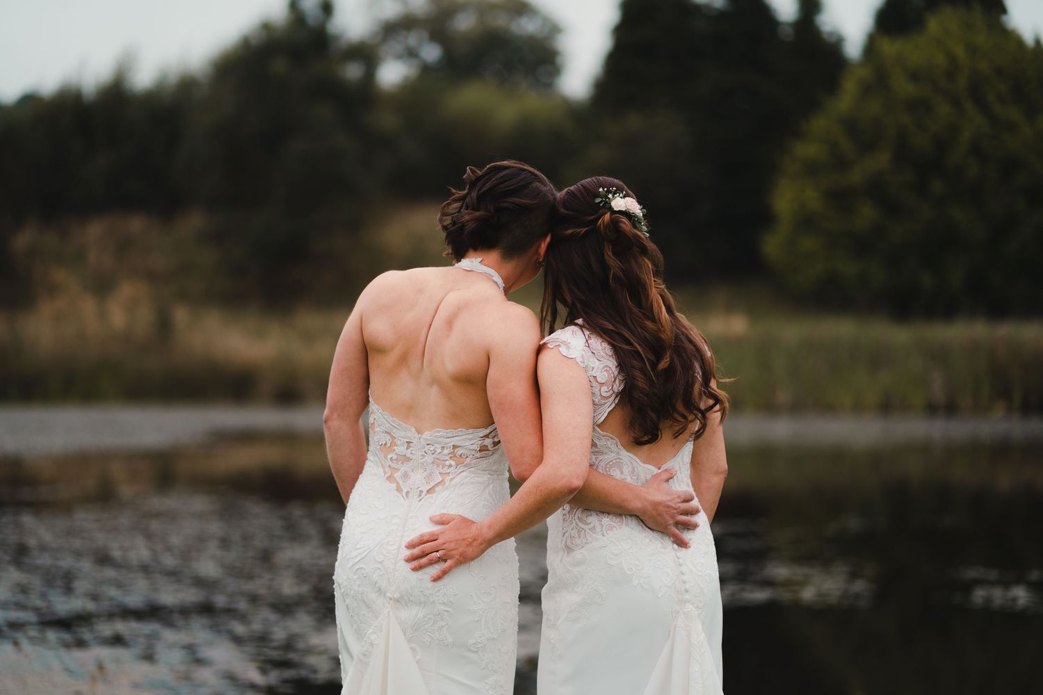 Two elegant brides in white lace wedding dresses stand next to each other on a lakeside dock at sunset.