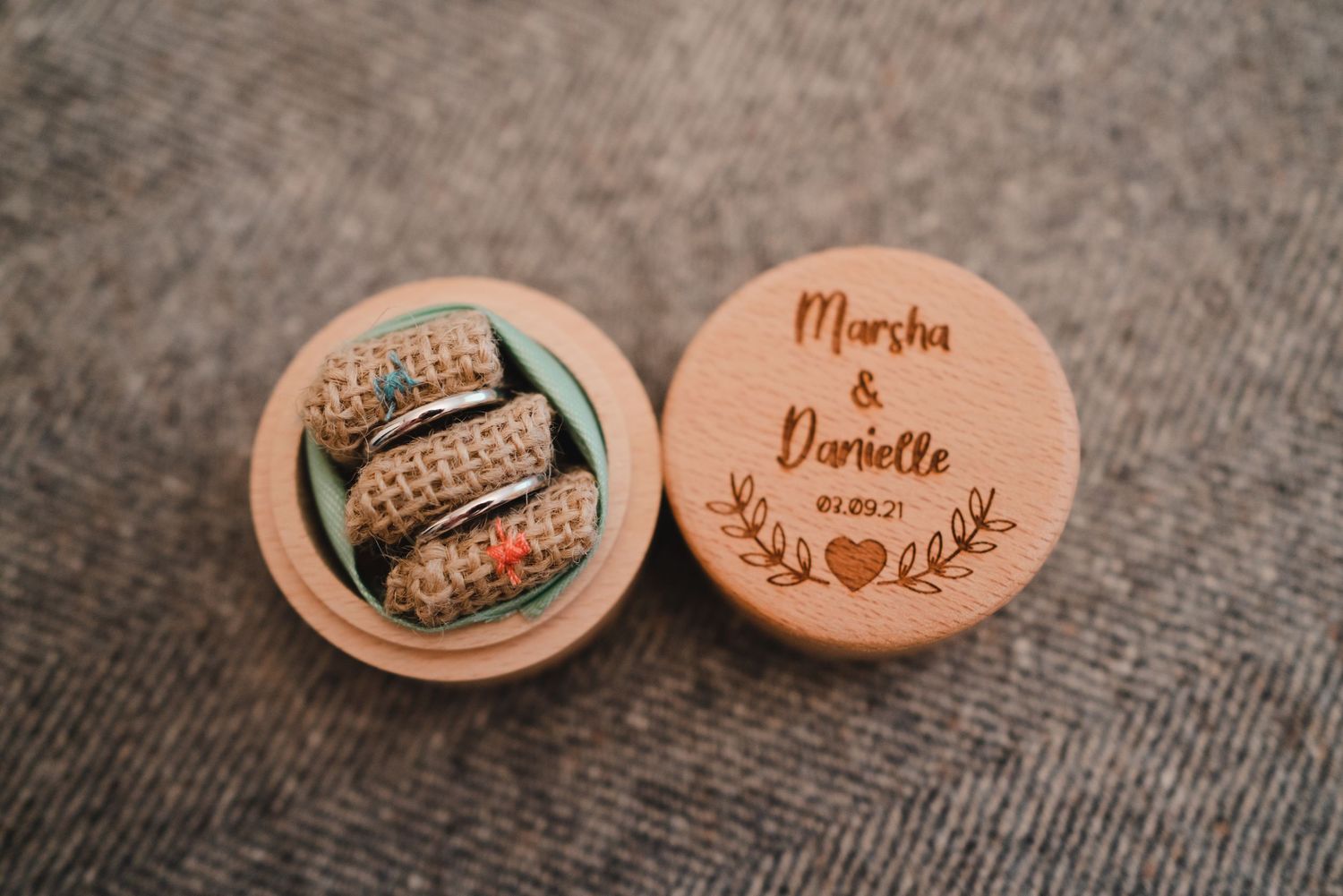Wooden ring boxes with engraved names displayed on textured gray fabric.
