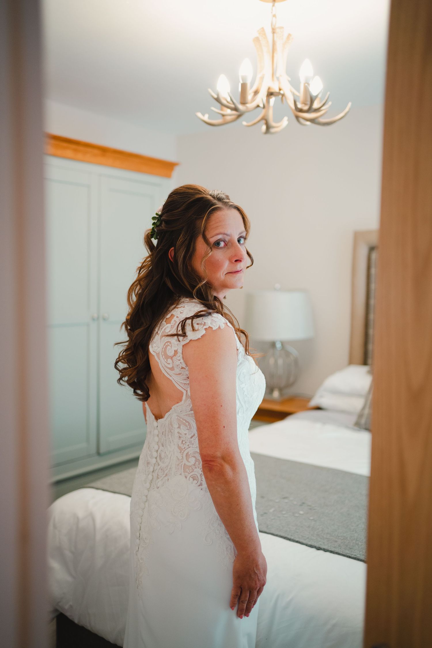 An elegant bride poses in a lace wedding dress in a bright bedroom with antler chandelier overhead.