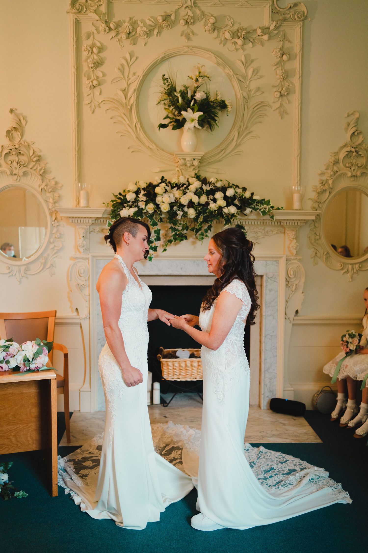 Two people in white wedding dresses share an intimate moment in an ornate room with classical decor.