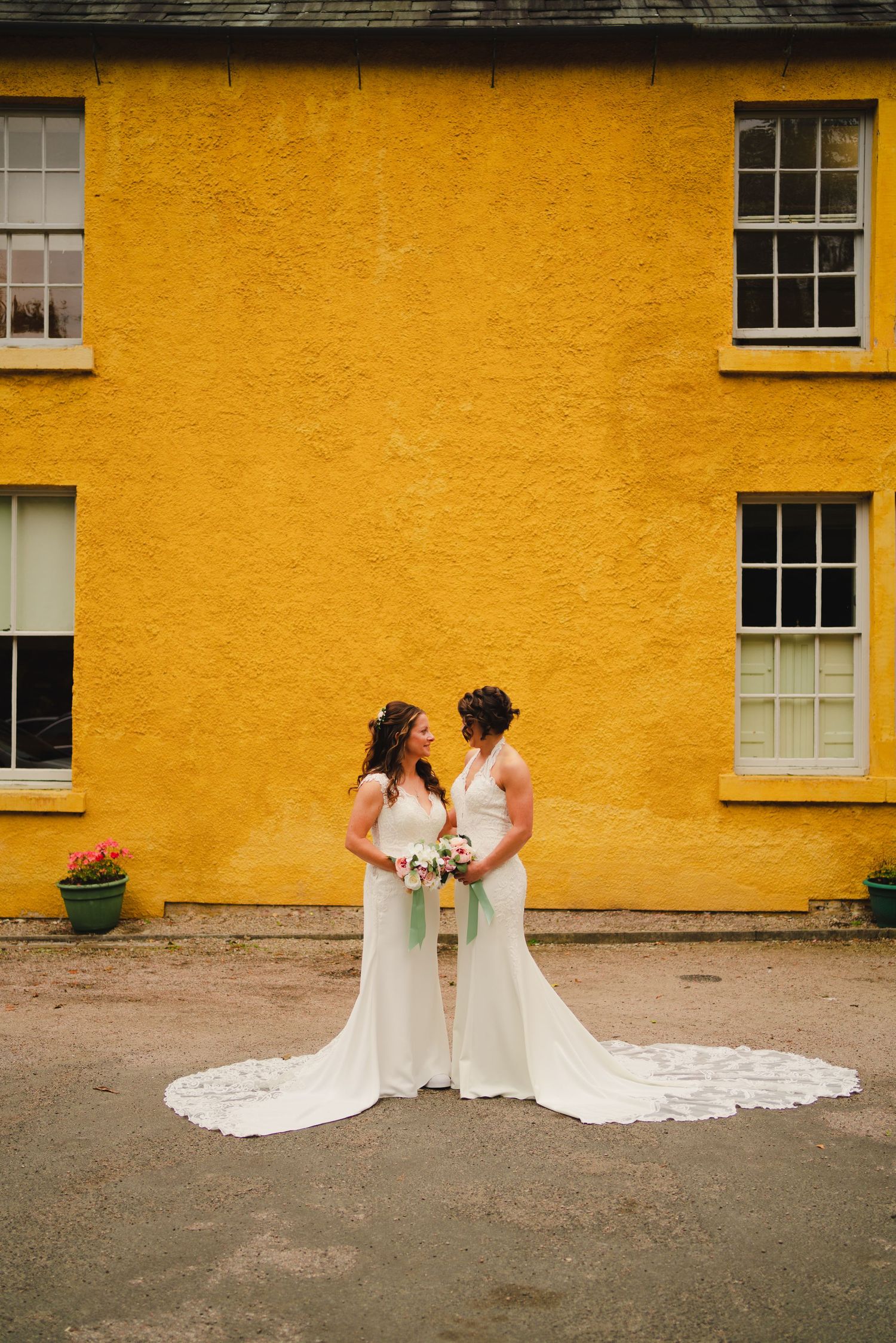 Two brides in long white wedding dresses with flowing trains stand together against a bright yellow wall.