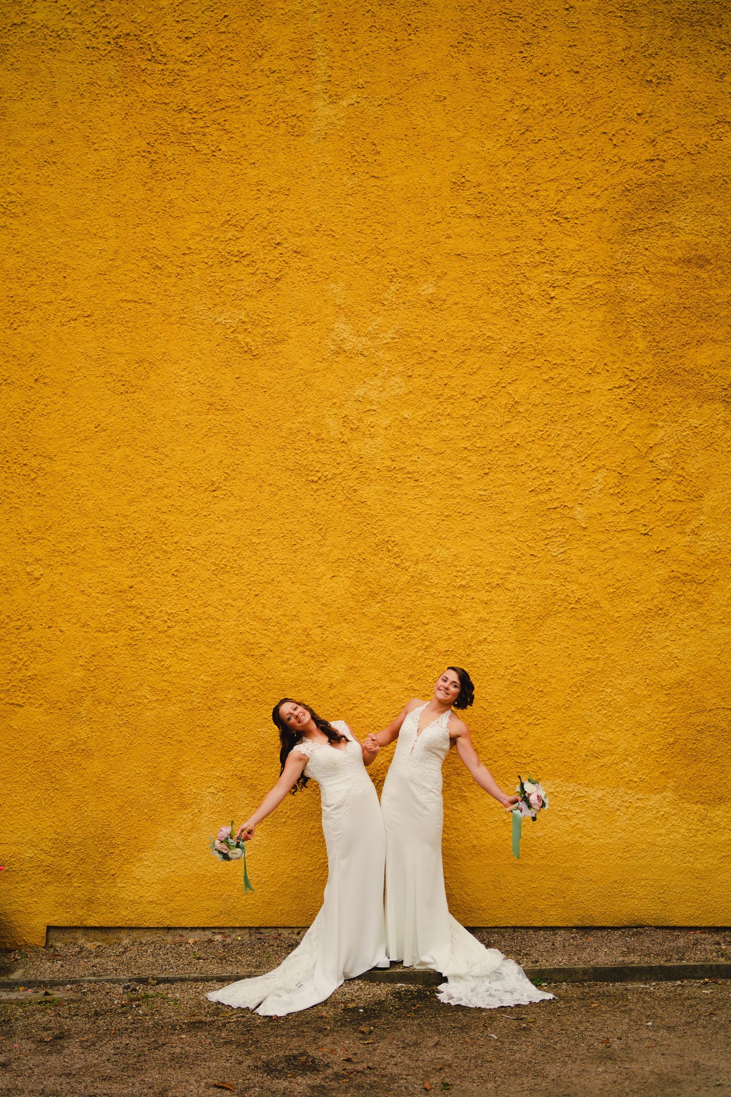 Two brides in white wedding dresses pose playfully against a vibrant yellow wall creating a striking contrast.