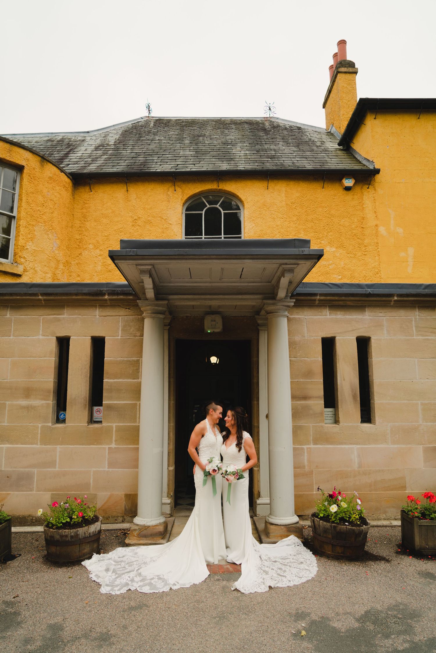 Elegant couple in white wedding dresses embrace in front of a yellow stone house with classical columns and arched doorway.