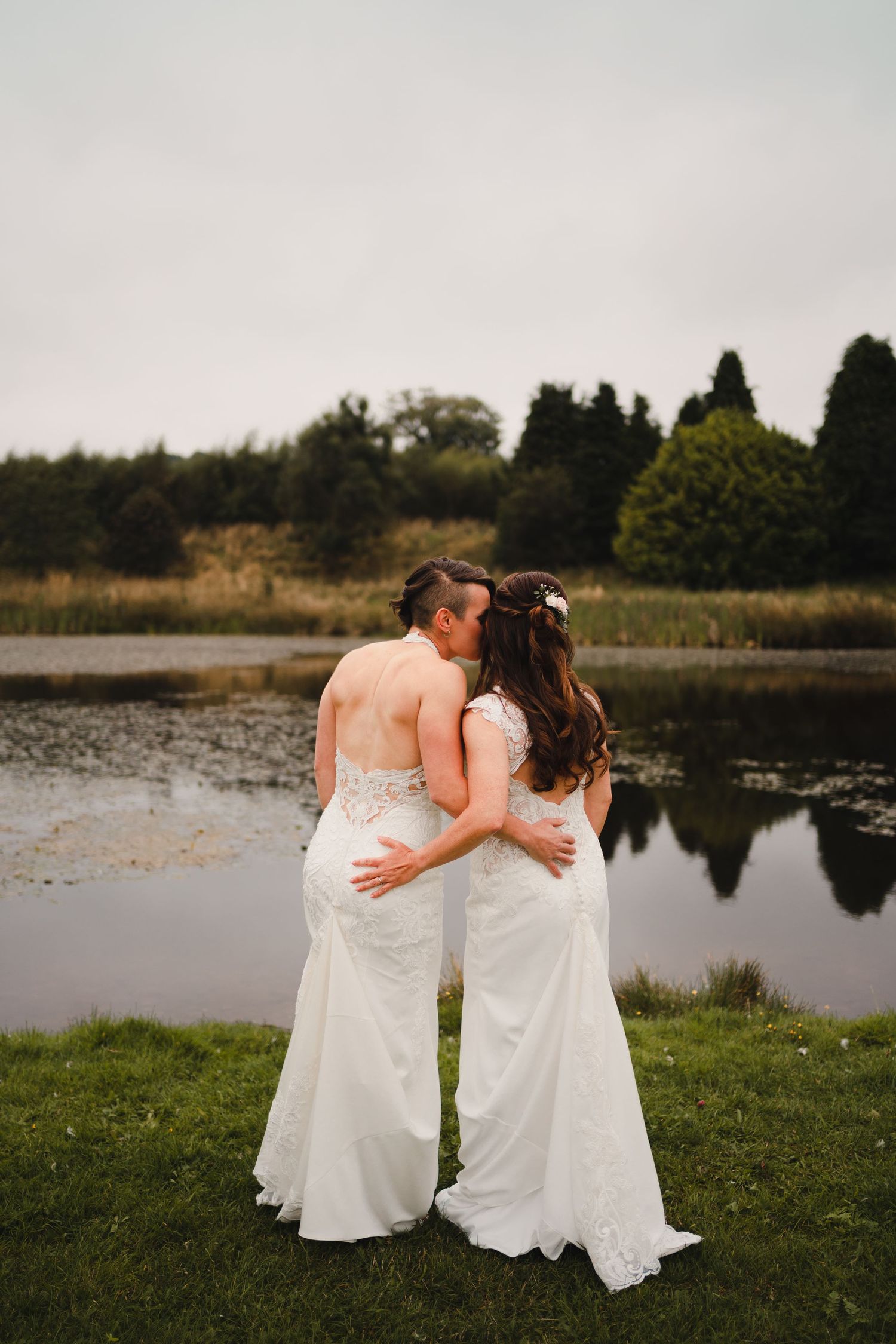 Two people in white dresses embrace while standing by a serene lake at sunset.