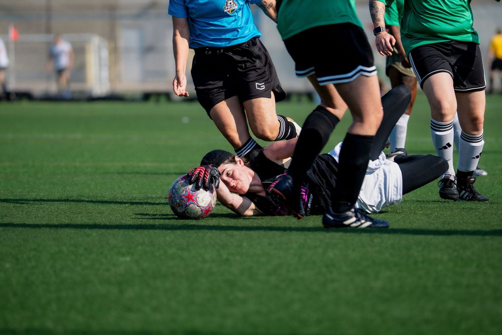 A soccer goalie laying on the ground with hand outstretched traps the ball to secure it at stop the drive