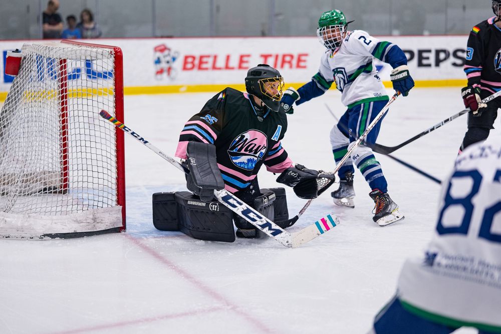 A hockey goalie kneels in front of of the goal to stop the players nearby in blue and white jerseys from scoring