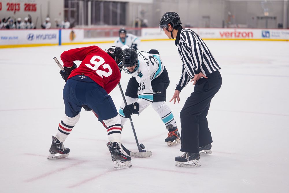 A hockey player in red and their opponent in a blue and white jersey fight for the puck that is dropped by the referee