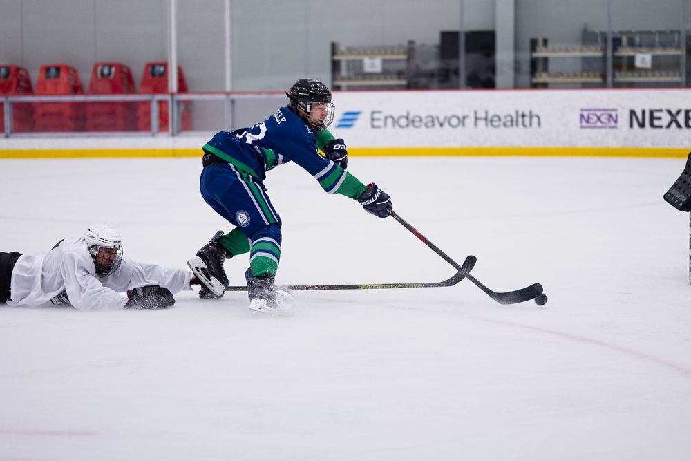 A hockey player in a blue and green jersey controls the puck in front of themselves while an opponent dives in attempt to recover