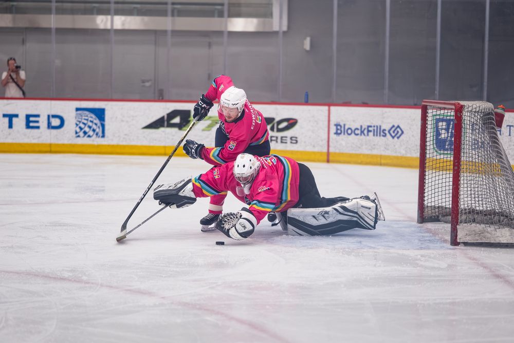 A hockey goalie and their teammate in pink jerseys make dives over each other to secure the puck near the net