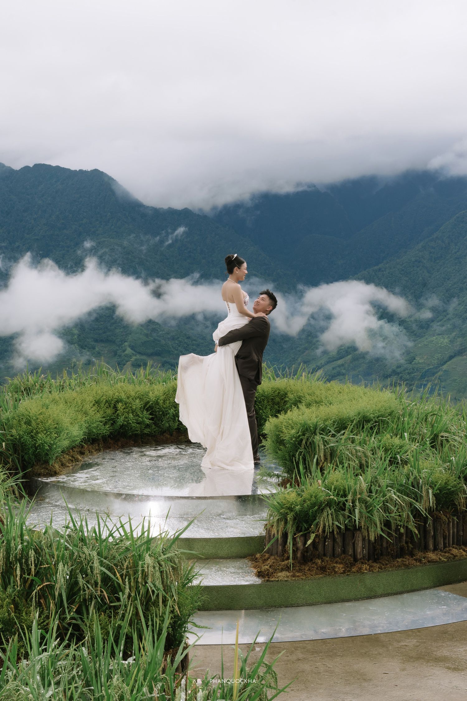 Bride and groom embrace on a circular stone platform surrounded by lush green rice terraces and misty mountain backdrop.