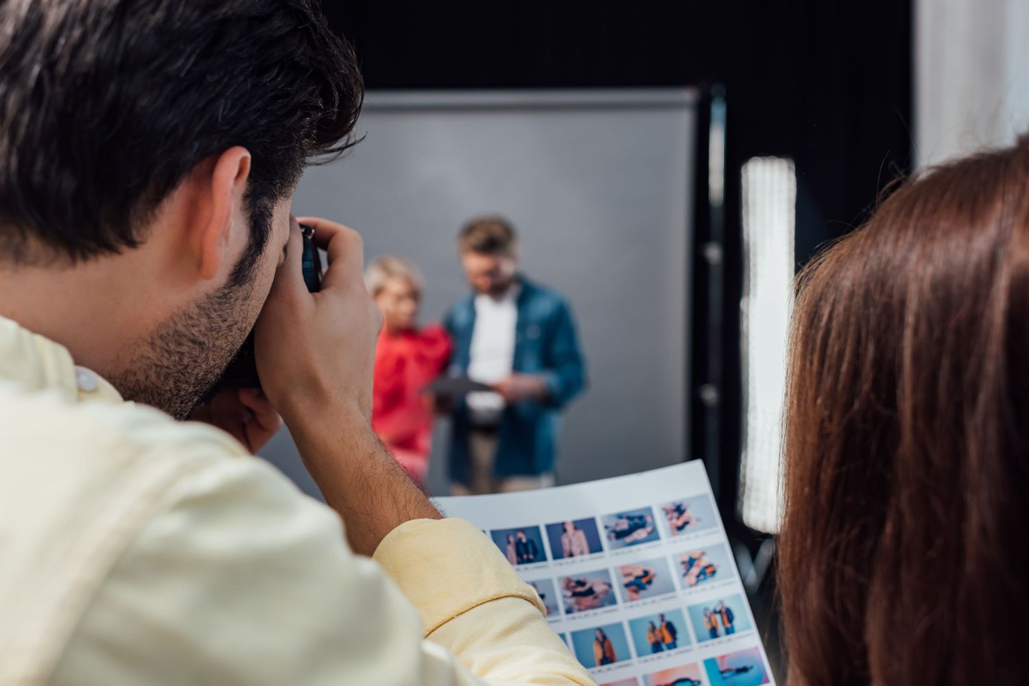 Photographer reviews photos on contact sheet while models wait in the background during fashion photoshoot.