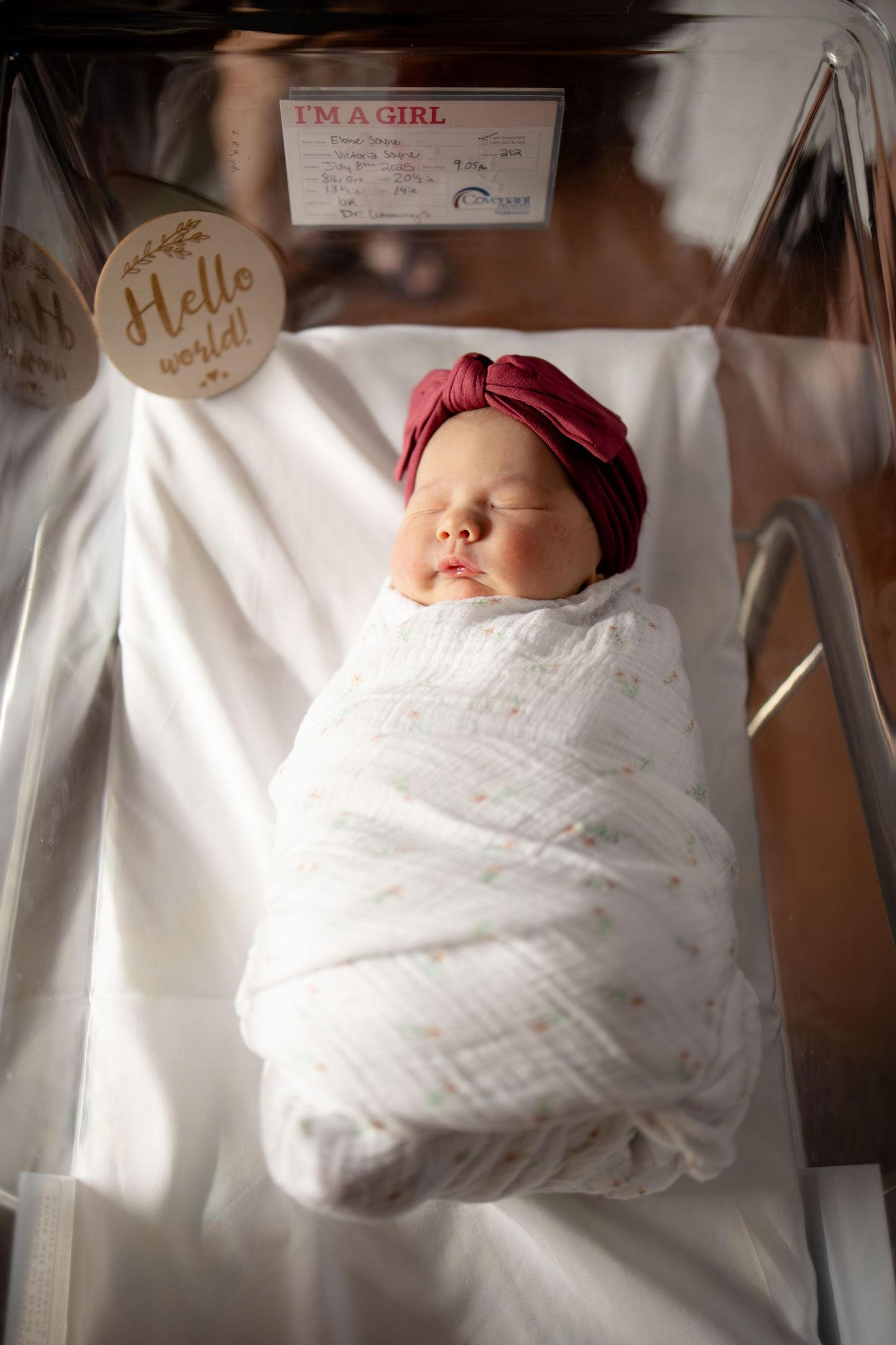 Swaddled newborn baby girl in burgundy turban hat sleeping in hospital bassinet with Hello World sign.