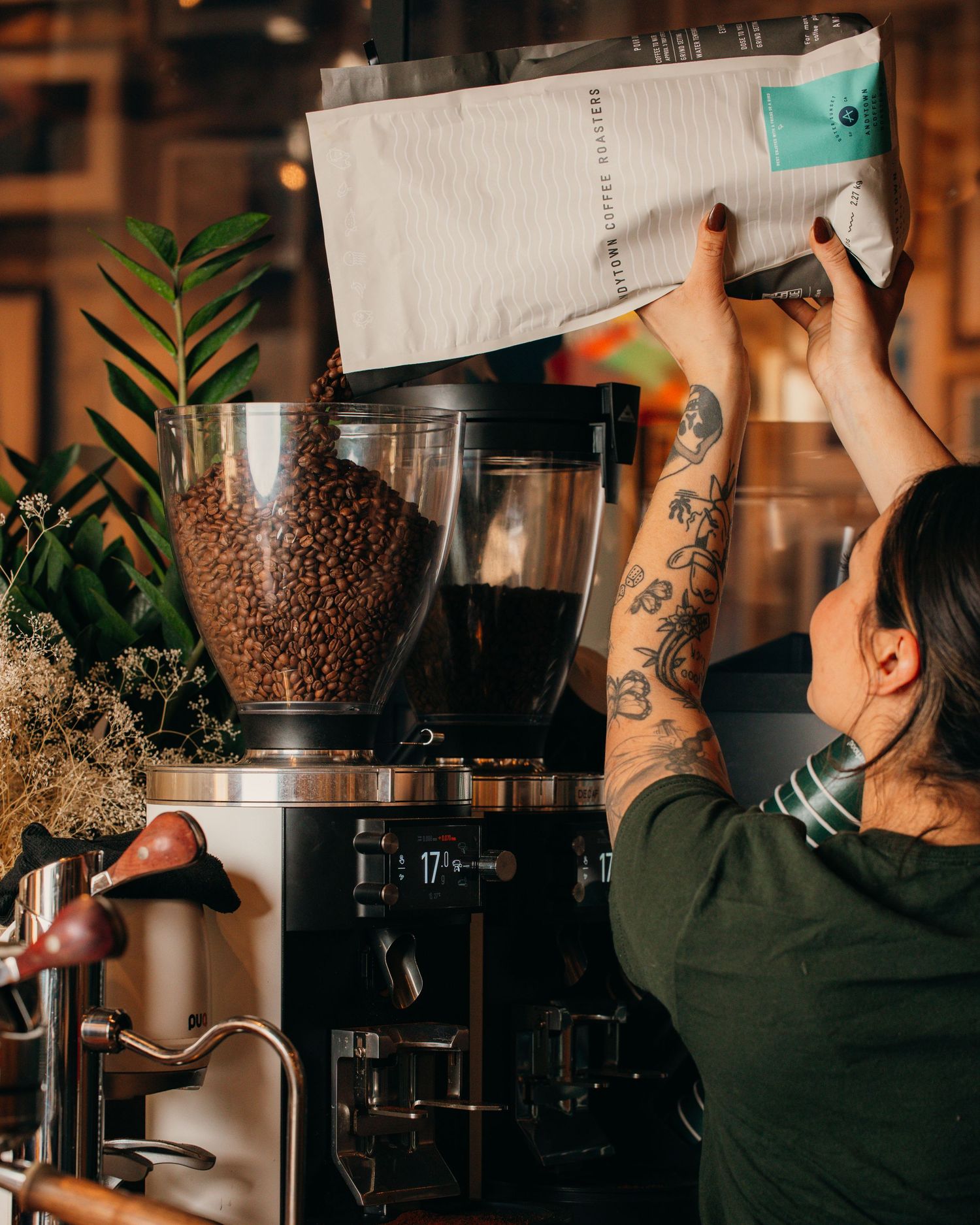 Barista with tattoos pouring coffee beans into commercial grinder at rustic coffee shop counter.