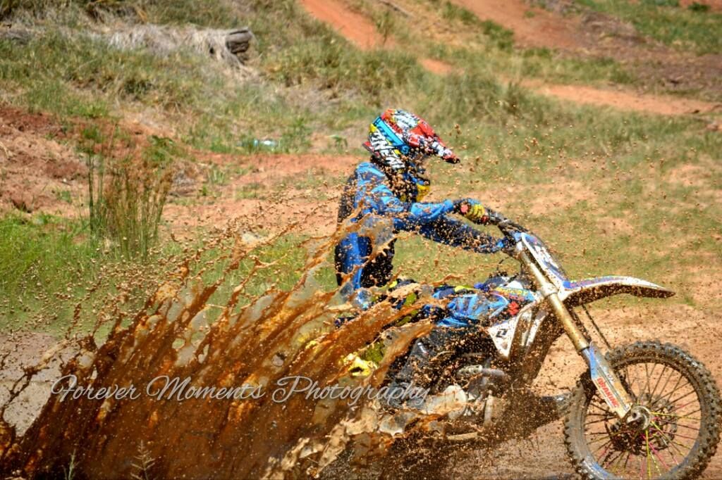 Dirt bike rider skillfully navigates a muddy motocross track with water spraying during an off-road competition.
